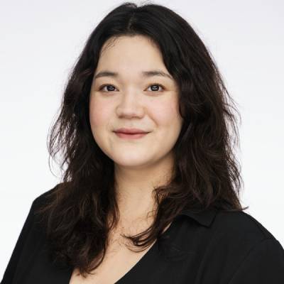 Person with medium-length, dark and slightly wavy hair, wearing a dark shirt, photographed against a light background.