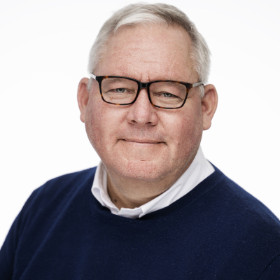 The picture shows a man with short grey hair, glasses and a calm expression. He is wearing a white shirt and a dark blue sweater.