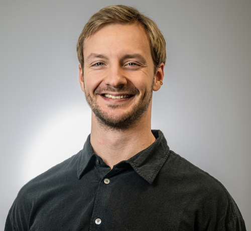 Man with blond hair combed to the side and light beard, wearing a dark shirt. The background is light gray.