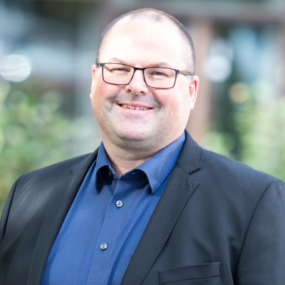 Portrait of a smiling man with glasses, a blue shirt and a dark suit jacket.