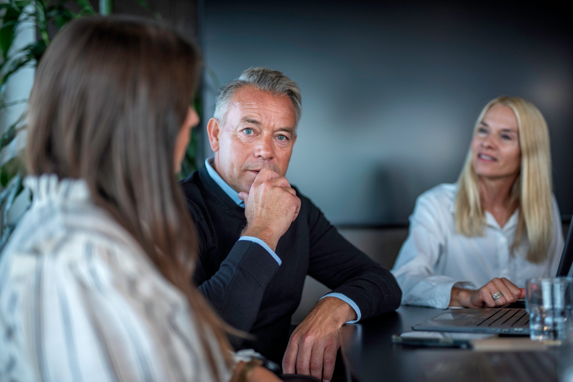 En mannlig og to kvinnelige kontorarbeidere sitter i et moderne møterom og har en samtale, mannen ser betenkt inn i kamera.