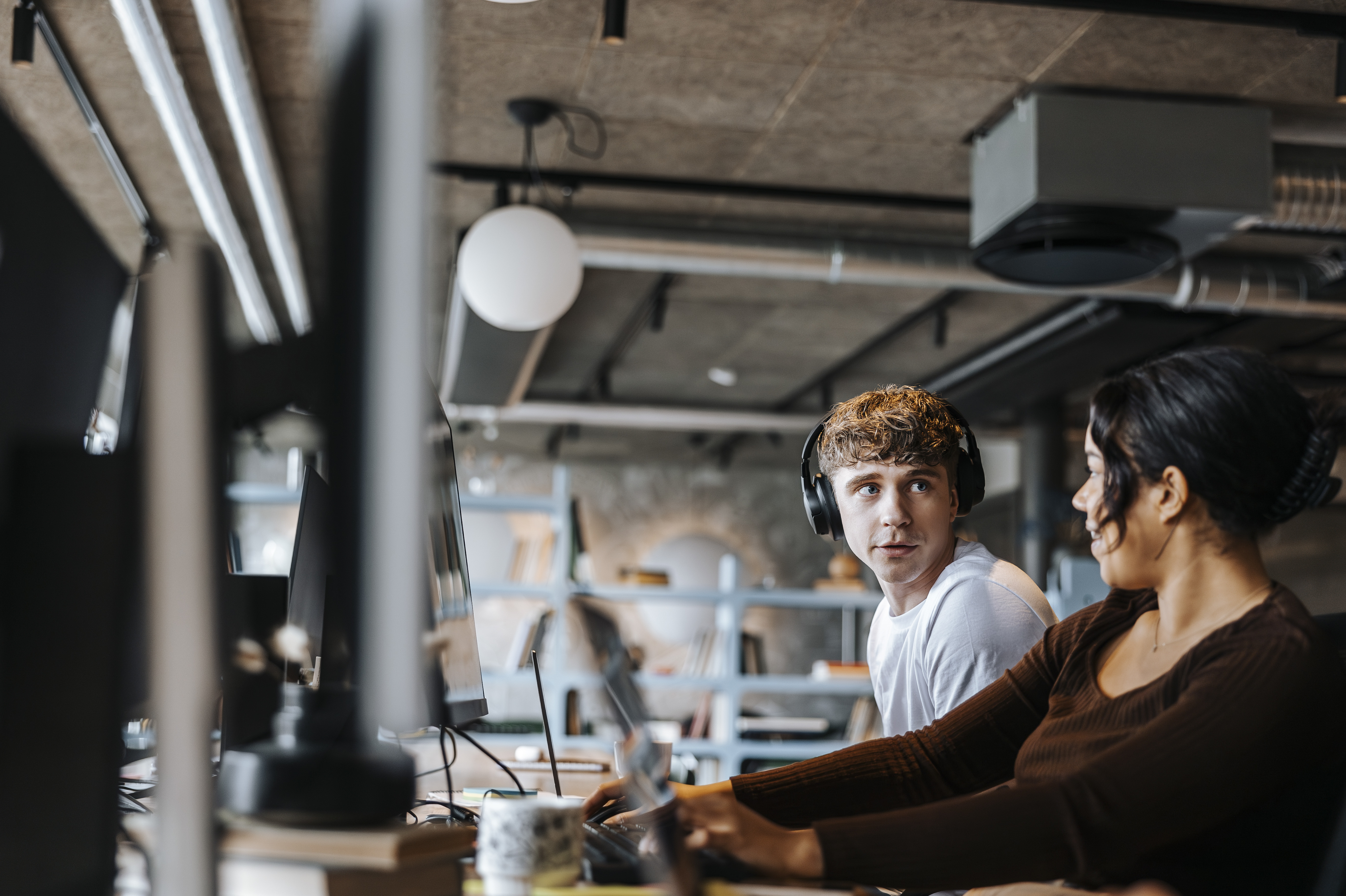 To unge ansatte, en kvinne og en mann, snakker sammen mens de sitter og jobber ved datamaskinene sine i et åpent kontorlandskap.