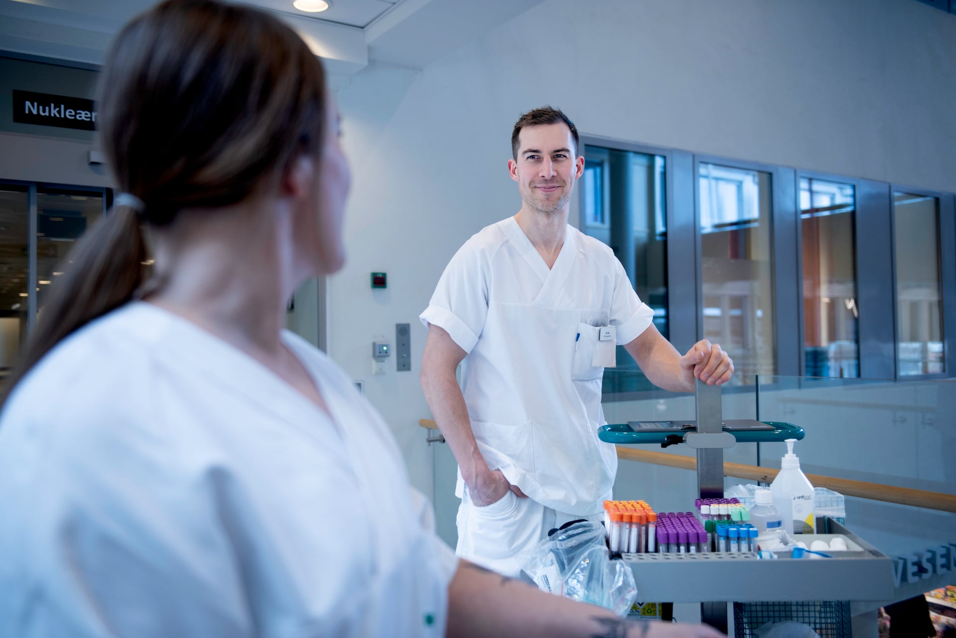 To uniformerte bioingeniører helsearbeider smiler til hverandre på sykehuset med en tralle med prøvetakingsutstyr 
