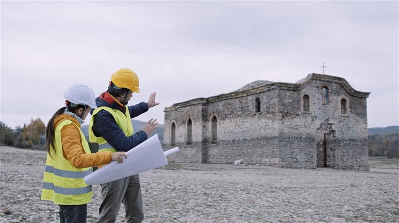 Two engineers in protective equipment discuss construction plans in front of an old building, symbolizing the importance of safety and good working conditions ensured through collective agreements in the building and construction industry.
