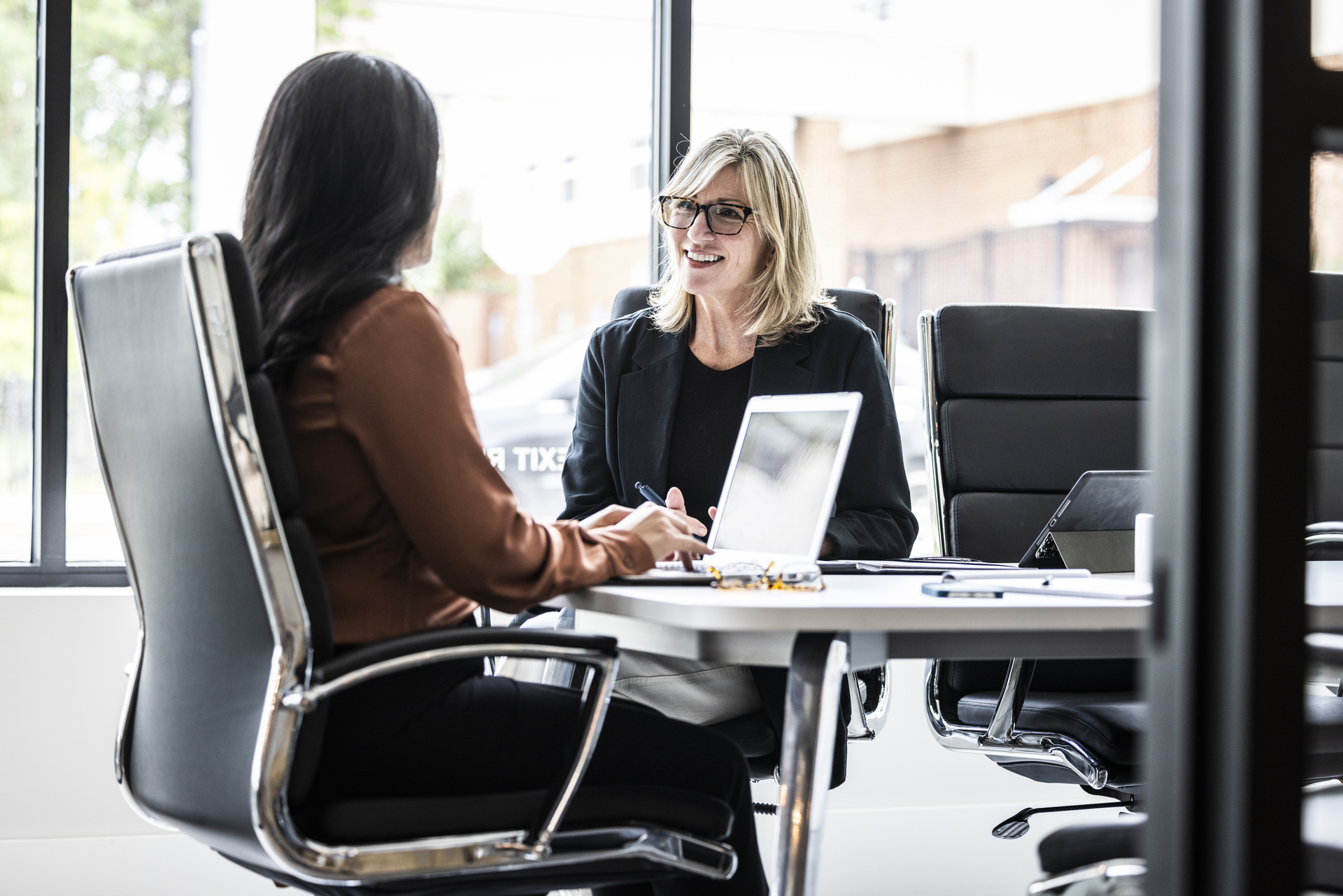 Two businesswomen sit in a modern conference room and discuss. One woman smiles and looks at the other, who is sitting with her back to her. A laptop is on the table between them. The picture can illustrate negotiations or cooperation related to collective agreements.