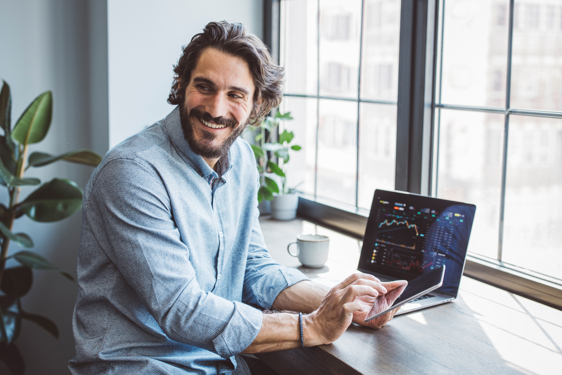 A smiling man sits at a table with a laptop and a tablet, looking out the window. The picture can illustrate flexibility in working life, which can be part of collective agreements.