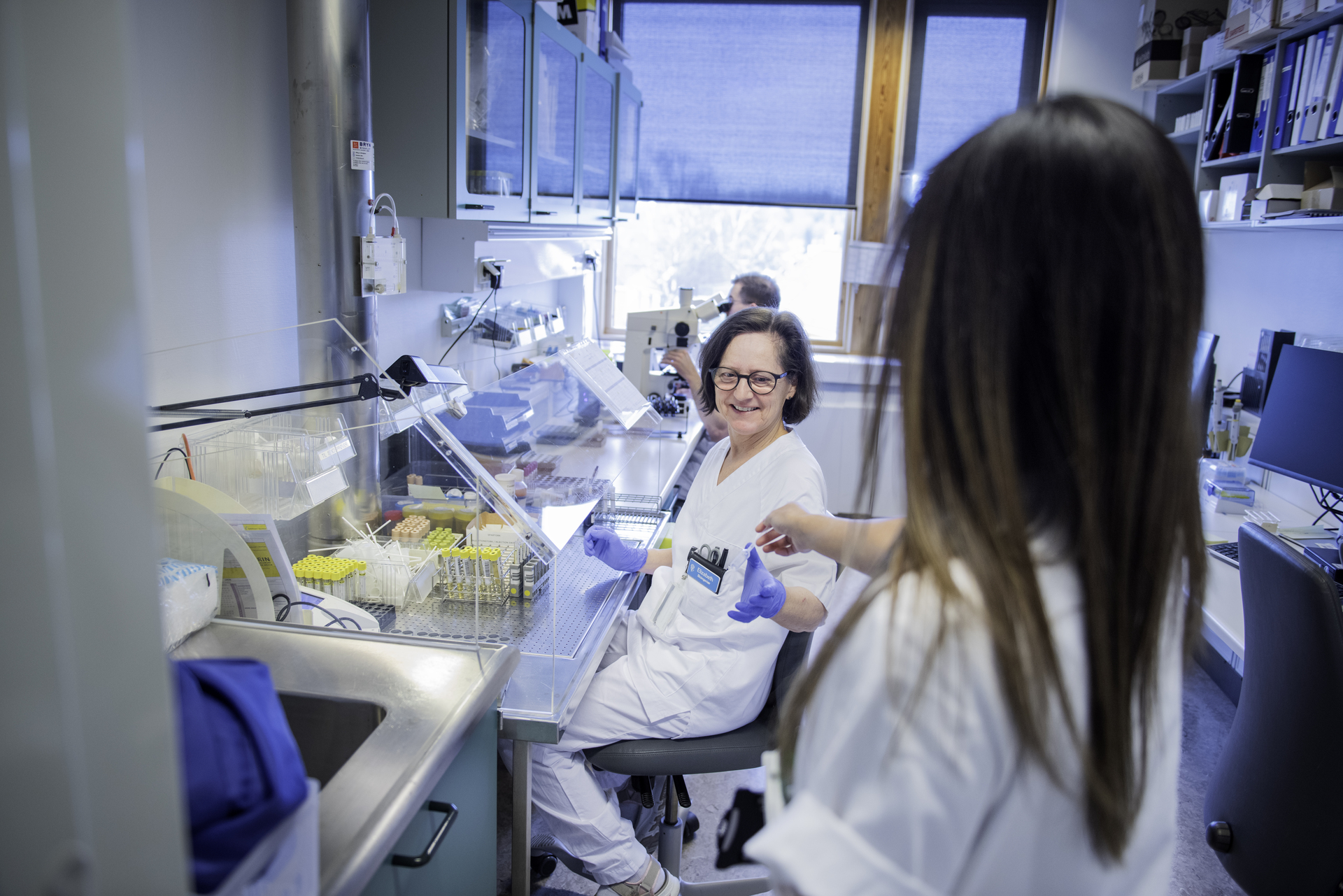 Two scientists or laboratory employees in white coats and gloves interact in a laboratory. The picture illustrates employees in the health sector who may be covered by a collective agreement