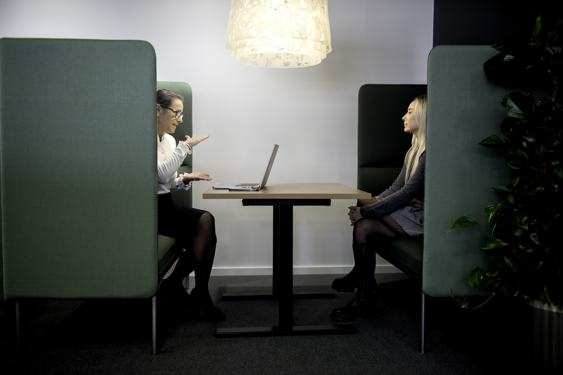 Two people are sitting in a modern meeting room with green screened cubicles, a table between them and a lamp above.