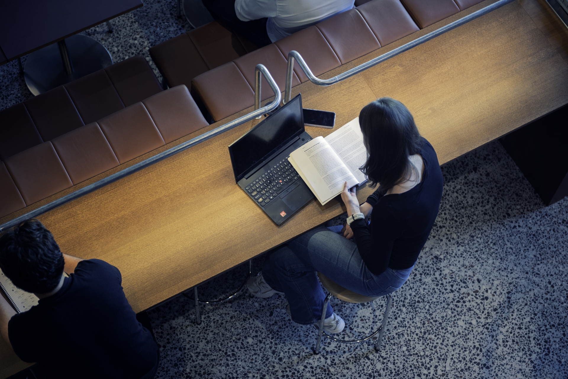 A person is sitting at a long table reading a book, with a laptop next to him in a quiet study environment.