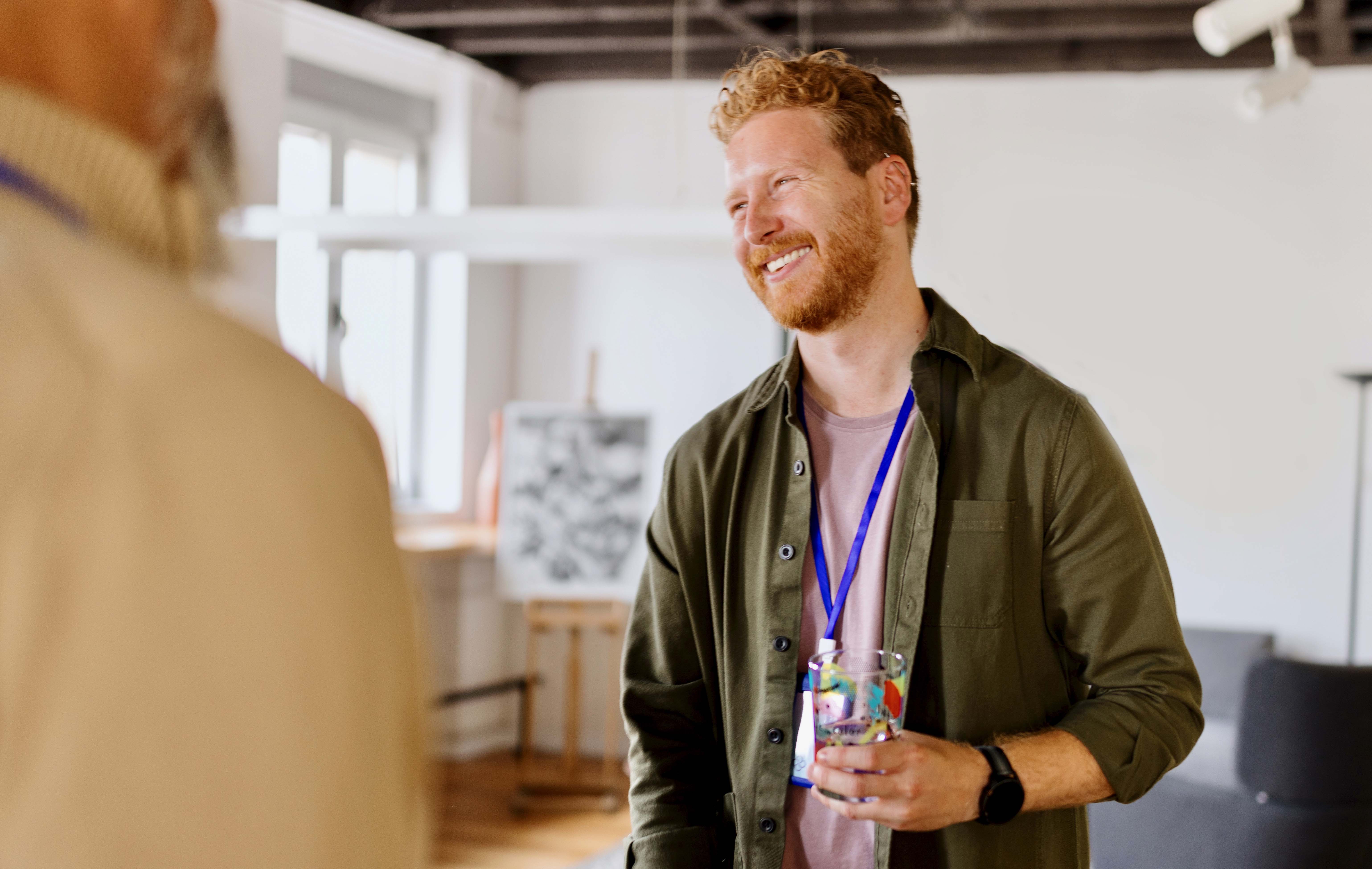 Smilende mann med skjegg holder et vannglass og snakker med noen vi bare ser ryggen på.
