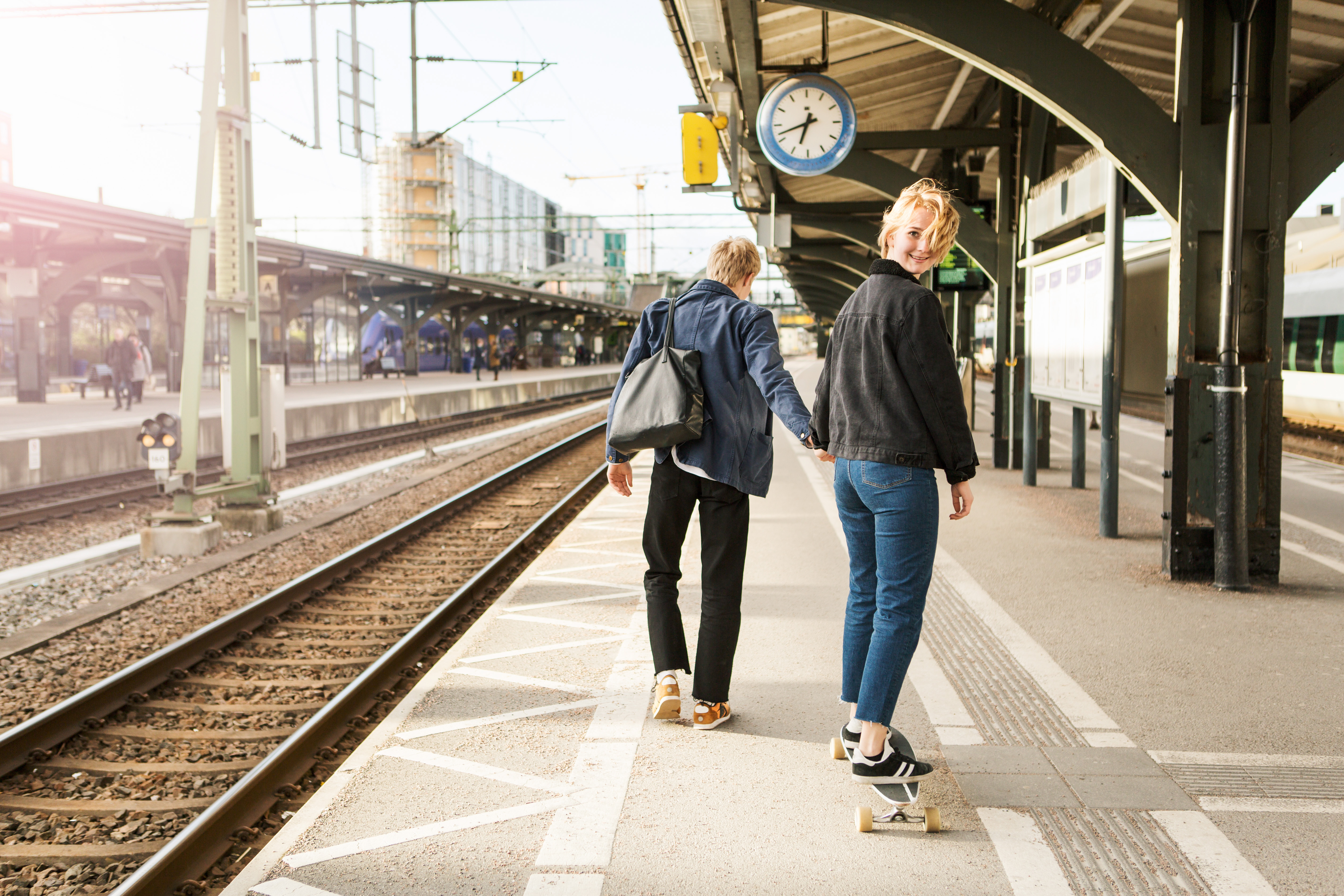 A young couple walks away from the camera at a train station, the woman has turned her head and looks at the camera with a smile. Photo: GettyImages